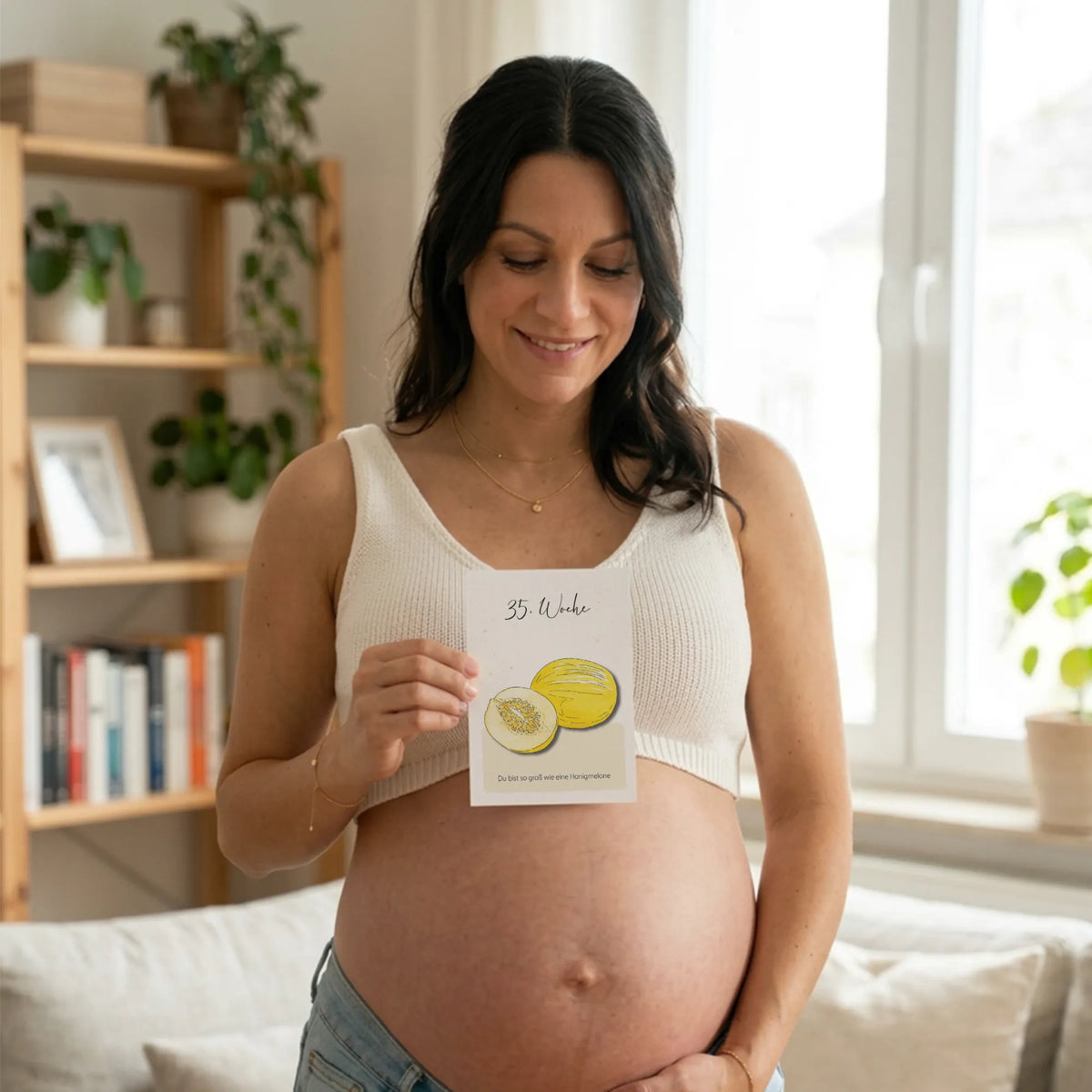 Frau mit Babybauch hält Meilensteinkarte mit SSW 35 und einer Melone vor ihrem runden Babybauch und lächelt.