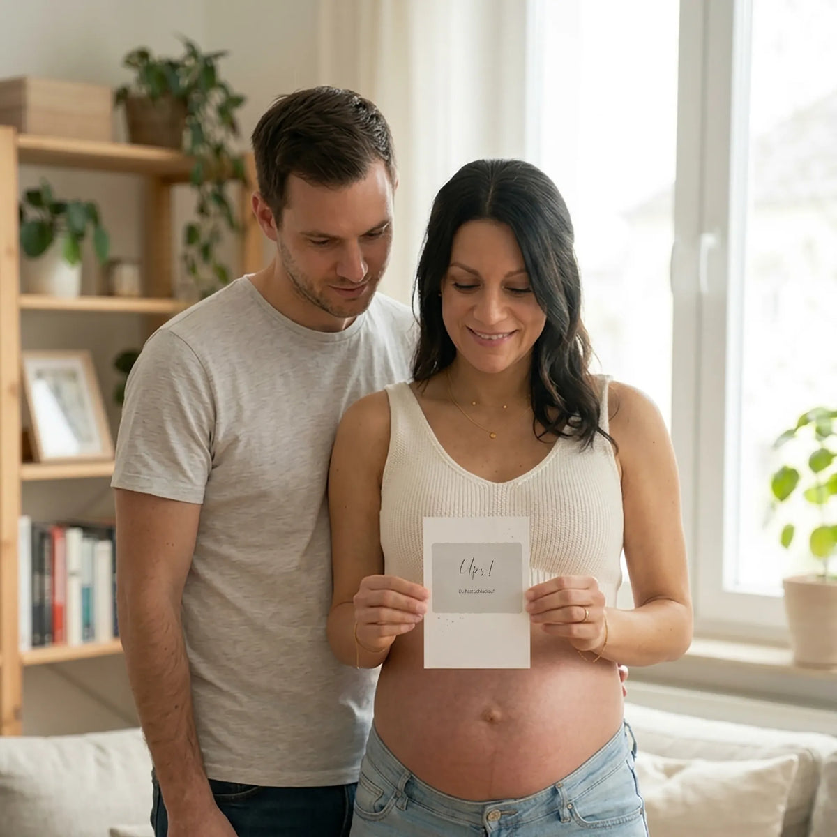 Pärchen mit Karte "Ups, du hast Schluckauf". Frau hält Karte vor ihren Babybauch, Mann steht hinter ihr.