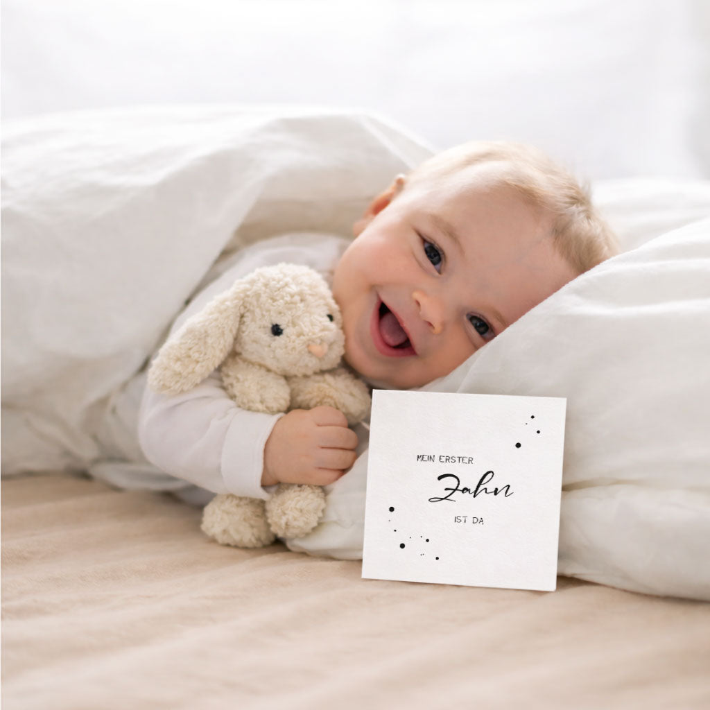 Baby im Bett mit Kuscheltier und Karte „Mein erster Zahn ist da“ – Meilensteinkarten fürs erste Jahr, um Fotos zu machen und besondere Momente als Erinnerung festzuhalten.