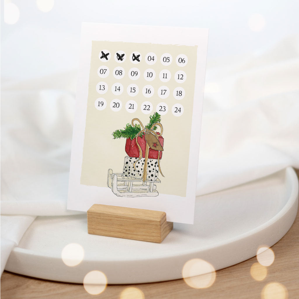 Adventskalender Countdown Karte mit Geschenken und Holzschlitten steht in Kartenhalter aus Holz vor Weihnachtsdeko