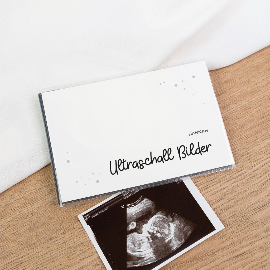 Ultraschallbilder Aufbewahrung Album in minimalistisch schwarz weiß: Einsteckalbum mit Schriftzug „Ultraschall Bilder“ und Name, davor ein Ultraschallfoto auf Holztisch.