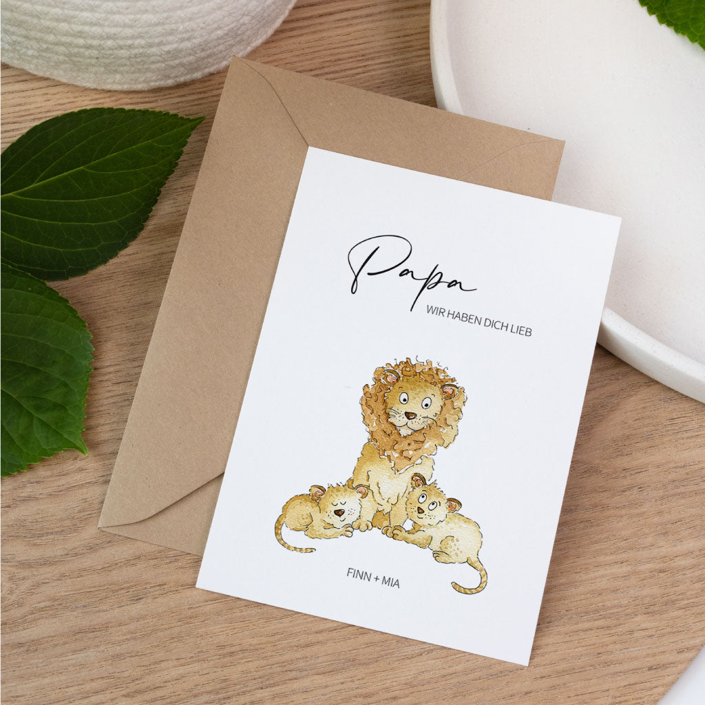 Vatertagskarte mit einem Papa Löwen und zwei Baby Löwen sowie der Aufschrift Papa und Wunschnamen auf einem Holztisch.
