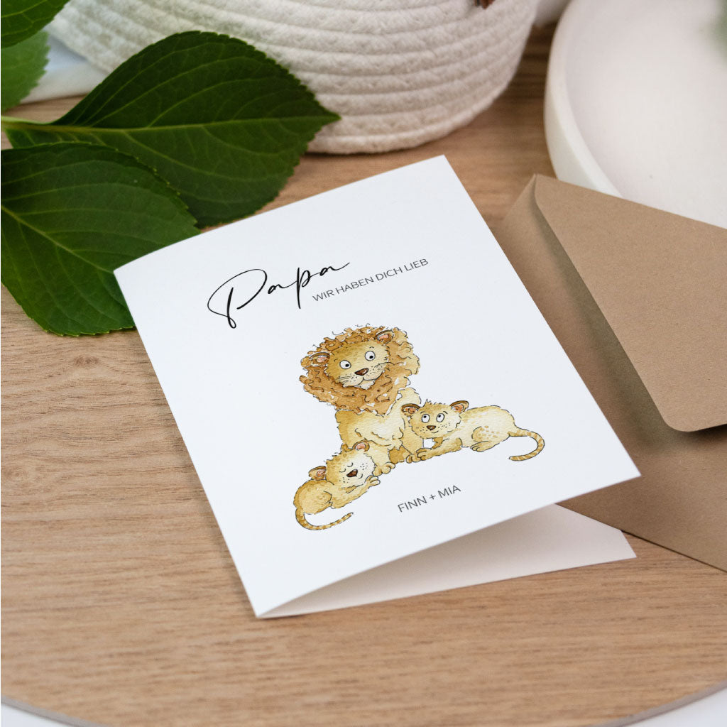 Aufgestellte Geburtstagskarte für den Papa mit Löwenfamilie und passendem Umschlag aus Kraftpapier neben grünen Blättern.