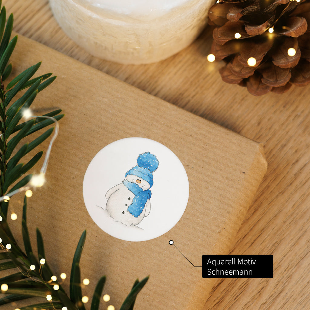 Runder Aufkleber mit Schneemann Aquarell Motiv auf braunem Verpackungspapier für festliche Geschenke.