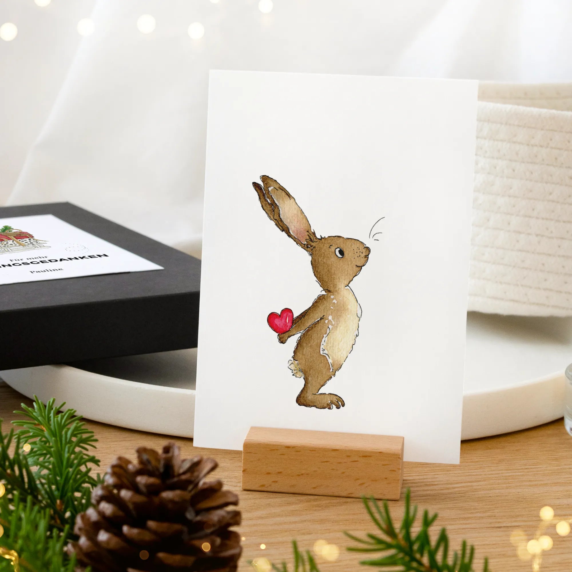 Aquarell Motiv Hase mit rotem Herz auf Karte im Holz-Kartenhalter, schwarze Geschenkbox im Hintergrund, Weihnachtsgeschenk Papeterie