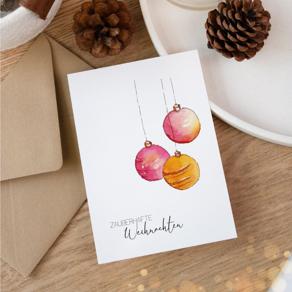 Handgemalte Weihnachtskarte mit drei Aquarell-Christbaumkugeln in Rot- und Orangetönen und dem Schriftzug „Zauberhafte Weihnachten“, liebevoll inszeniert mit Naturdeko.