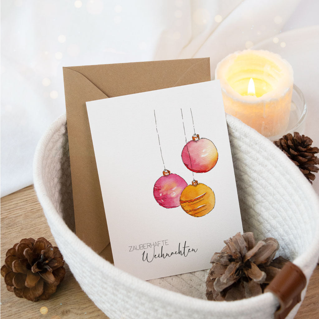 Besondere Klappkarte mit Aquarellmotiv aus drei hängenden Christbaumkugeln und weihnachtlichem Gruß, stimmungsvoll im Stoffkorb mit Kerze und Tannenzapfen präsentiert.
