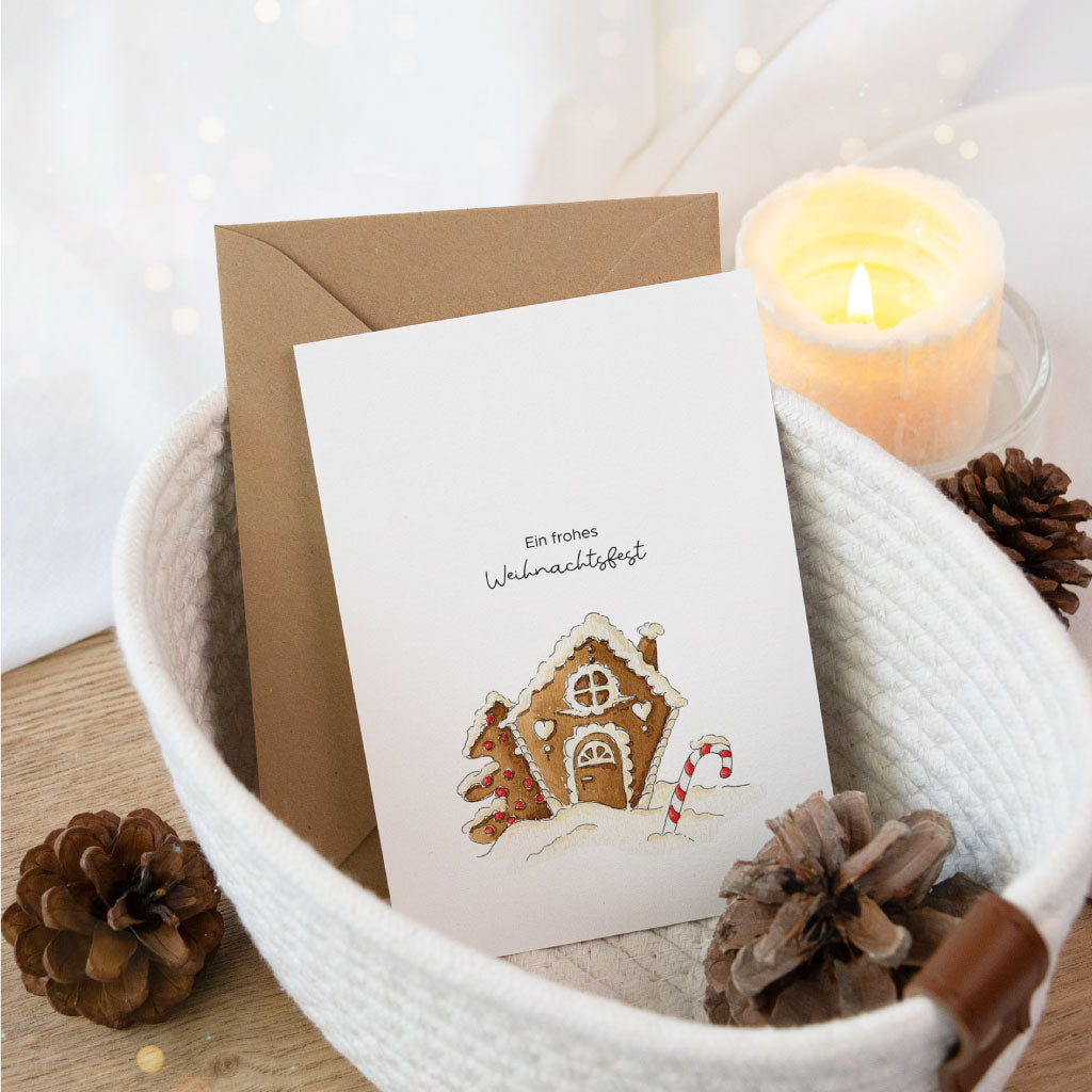 Weihnachtskarte mit Aquarellmotiv eines Lebkuchenhauses, stimmungsvoll arrangiert mit Deko, Tannenzapfen und Kerze – perfekt als Postkarte oder für die Geschenkverpackung zu Weihnachten.