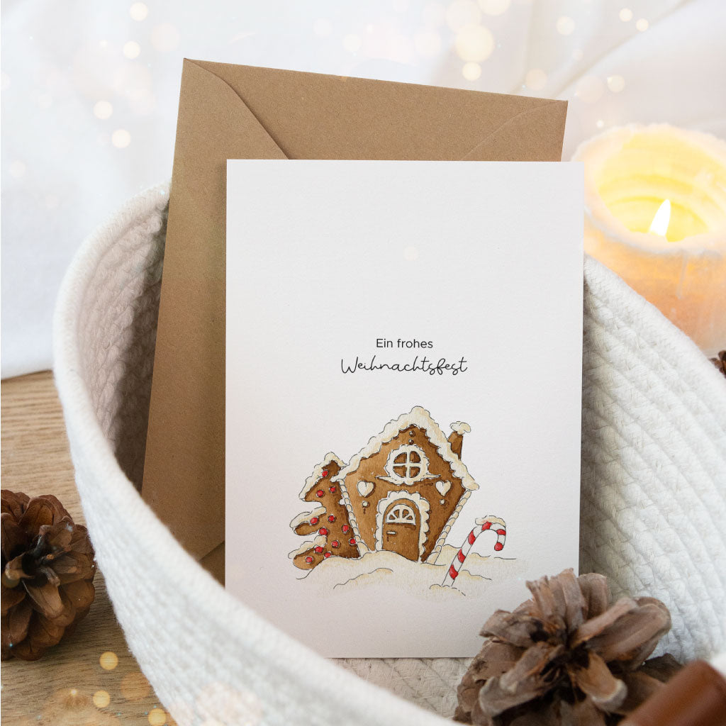 Postkarte mit handgemaltem Aquarell-Lebkuchenhaus und dem Schriftzug „Ein frohes Weihnachtsfest“, liebevoll dekoriert mit Kerzenschein und Naturmaterialien.