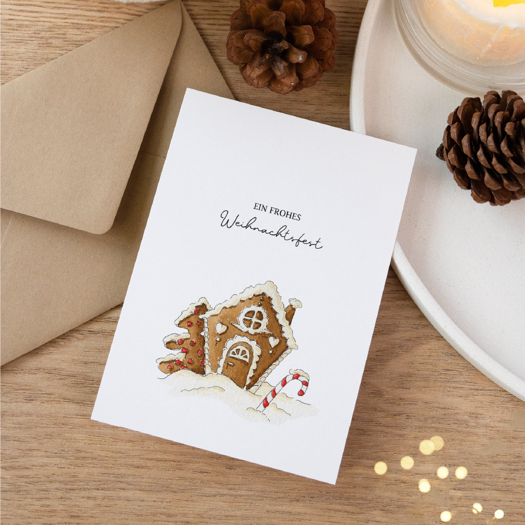 Postkarte mit handgemaltem Lebkuchenhaus Aquarell Motiv auf einem Holztisch mit Tannenzapfen.