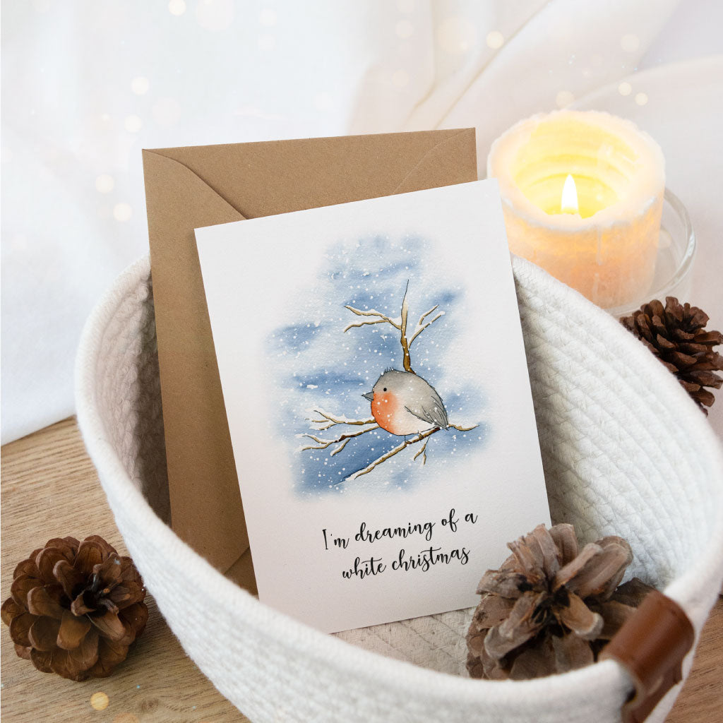 Weihnachtskarte mit handgemaltem Aquarell eines Rotkehlchens auf verschneitem Ast, dazu der Spruch „I'm dreaming of a white Christmas“, stilvoll arrangiert in einem winterlichen Geschenkkorb.