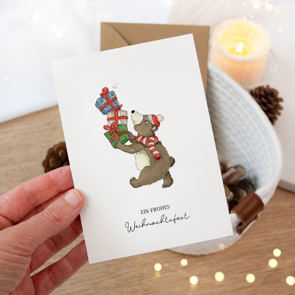 Eine Hand hält die weiße Klappkarte mit weihnachtlichem Bär Aquarell Motiv vor einem festlichen Korb.