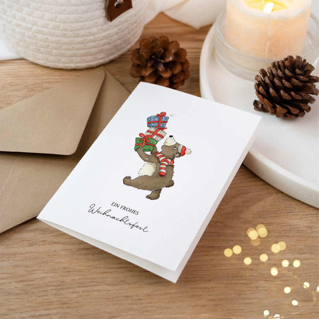Aufgestellte Weihnachtskarte mit Bär der bunte Geschenke trägt neben Tannenzapfen und Kerzenschein.