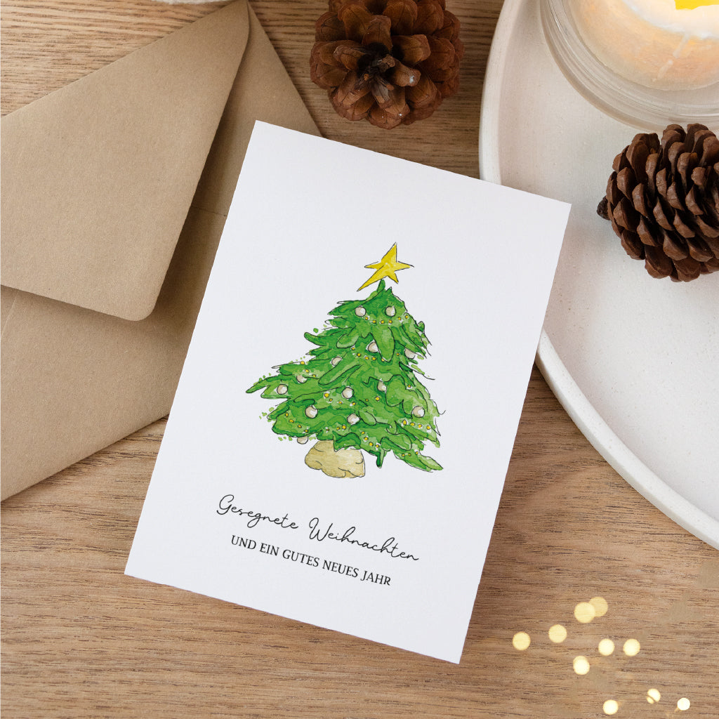 Festliche Weihnachtskarte mit Christbaum Aquarell Motiv liegend auf Holzhintergrund neben Tannenzapfen und Umschlag.