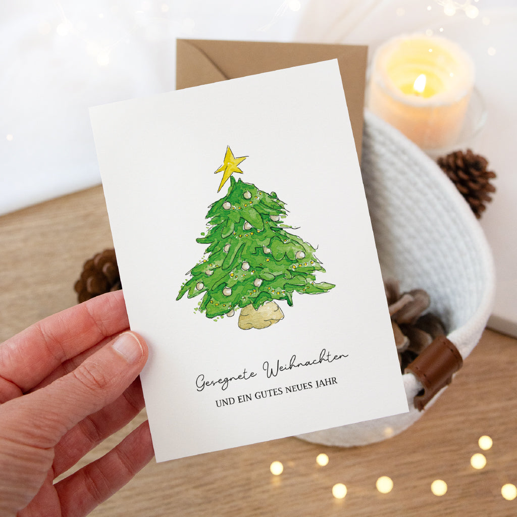 Hand hält eine festliche Klappkarte mit grünem Tannenbaum Aquarell Motiv vor einem weißen Korb mit Tannenzapfen.