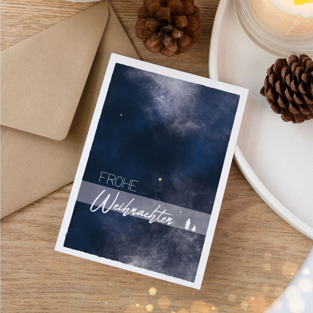 Besondere Weihnachtskarte mit handgemaltem Aquarellhimmel in Dunkelblau und dem Schriftzug „Frohe Weihnachten“, liebevoll arrangiert mit Umschlag, Kerze und Tannenzapfen.