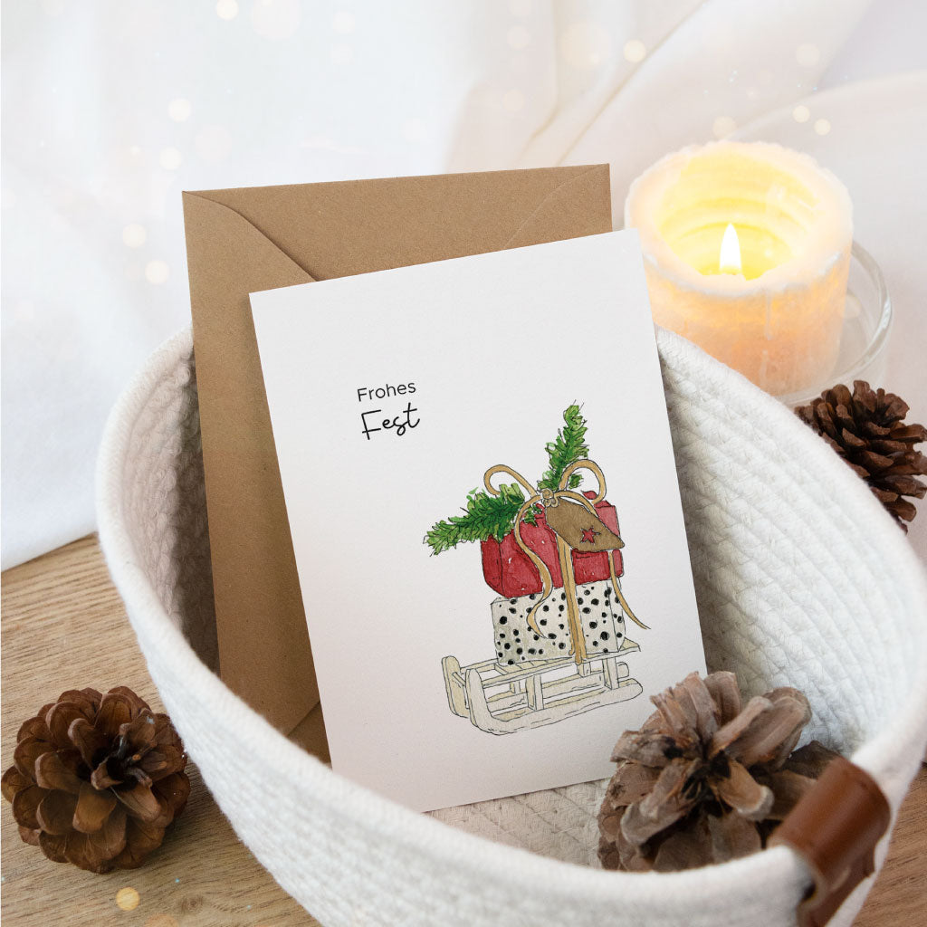 Weihnachtskarte mit handgemaltem Aquarell eines Schlittens voller Weihnachtsgeschenke und dem Spruch „Frohes Fest“, dekorativ präsentiert mit Kerze und Umschlag.