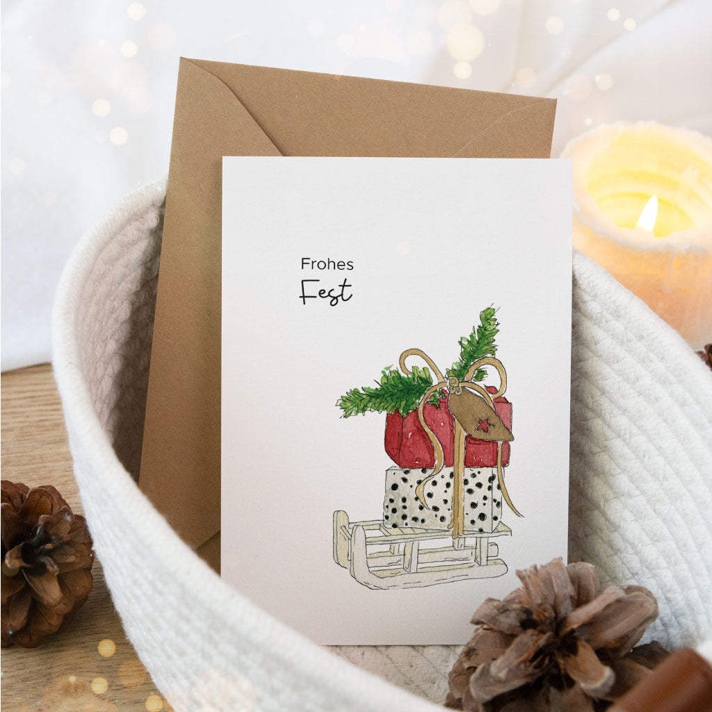Postkarte mit Aquarellillustration eines liebevoll gepackten Schlittens und Schriftzug „Frohes Fest“, perfekt zum Verschenken oder Weihnachtsgrüße schreiben.