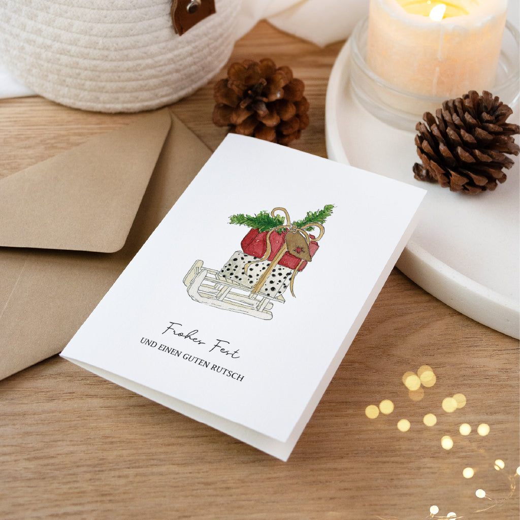 Klappkarte für Weihnachtsgrüße mit handgemaltem Schlitten Aquarell Motiv dekoriert mit Tannenzapfen auf Holz.