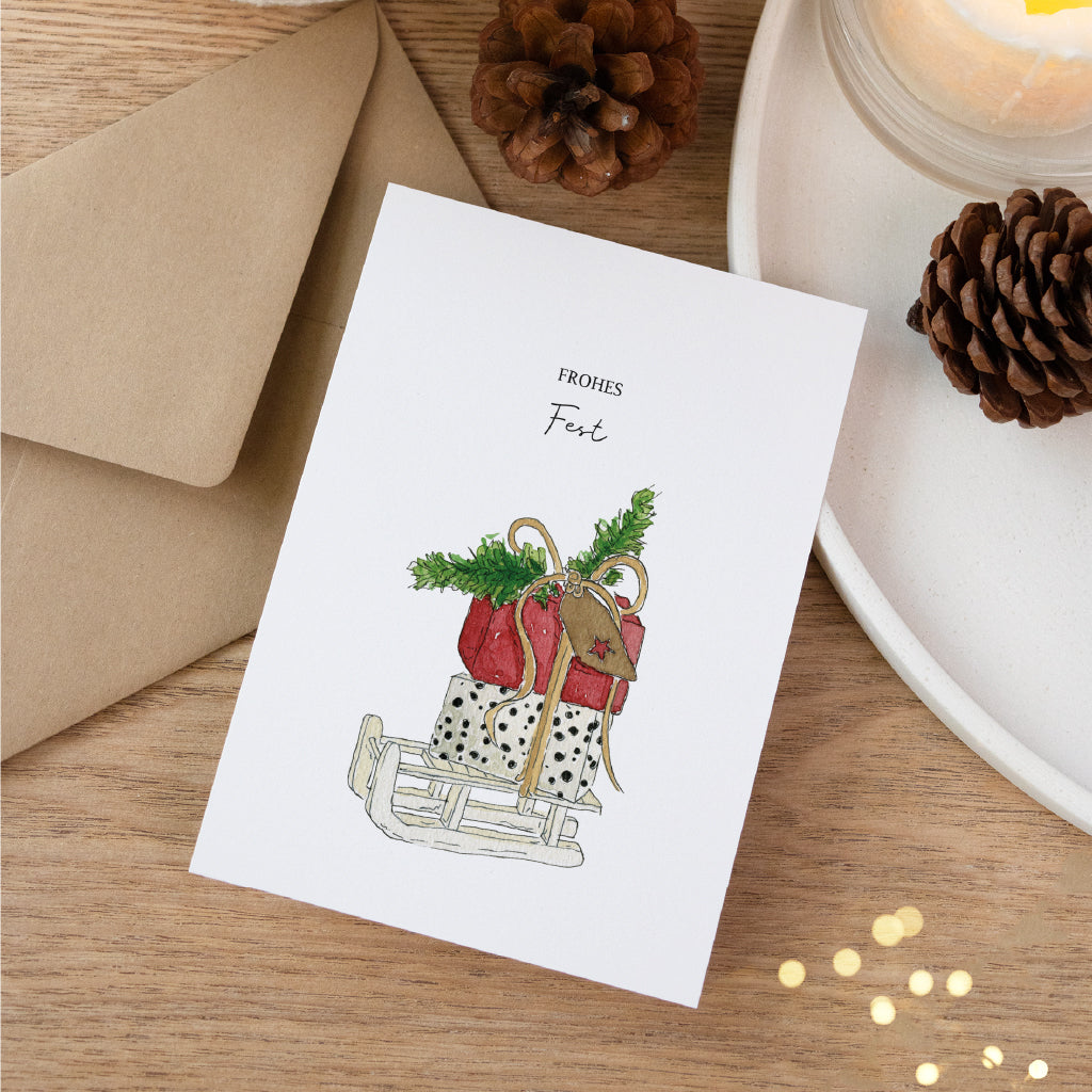 Handgemaltes Geschenke Aquarell Motiv auf Postkarte für Weihnachten dekoriert auf weißem Tablett mit Tannenzapfen.