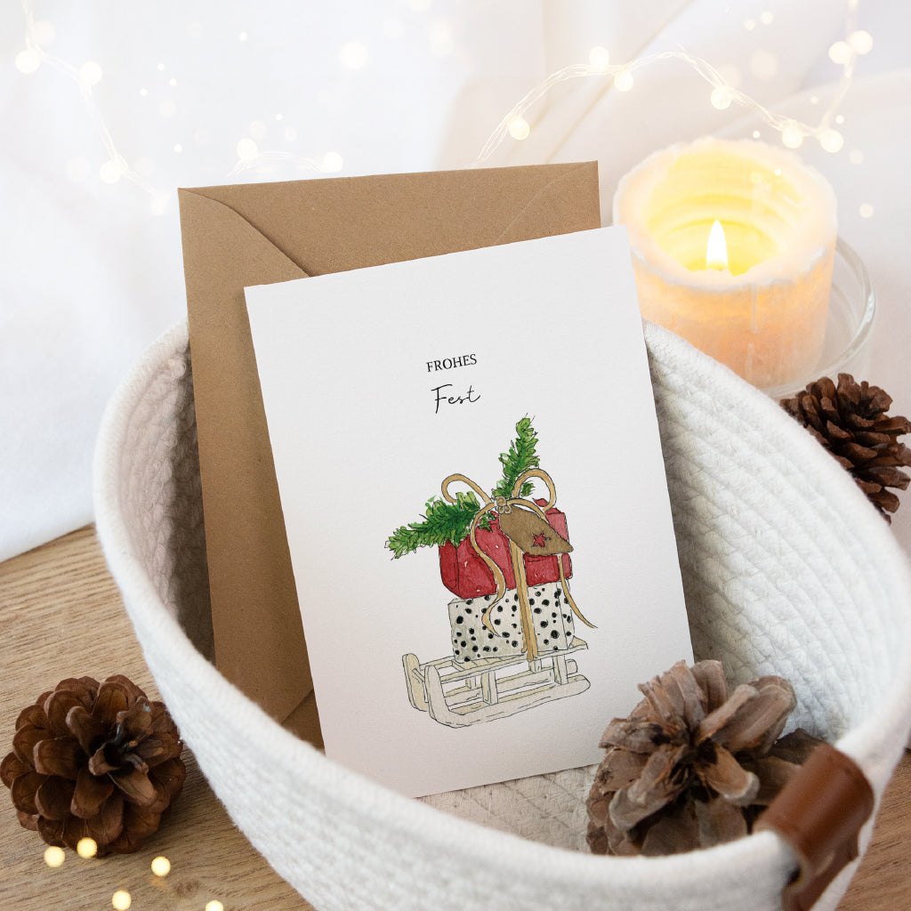 Stimmungsvolle Postkarte für die Weihnachtspost mit Schlitten und Geschenken im Korb bei gemütlichem Kerzenschein.