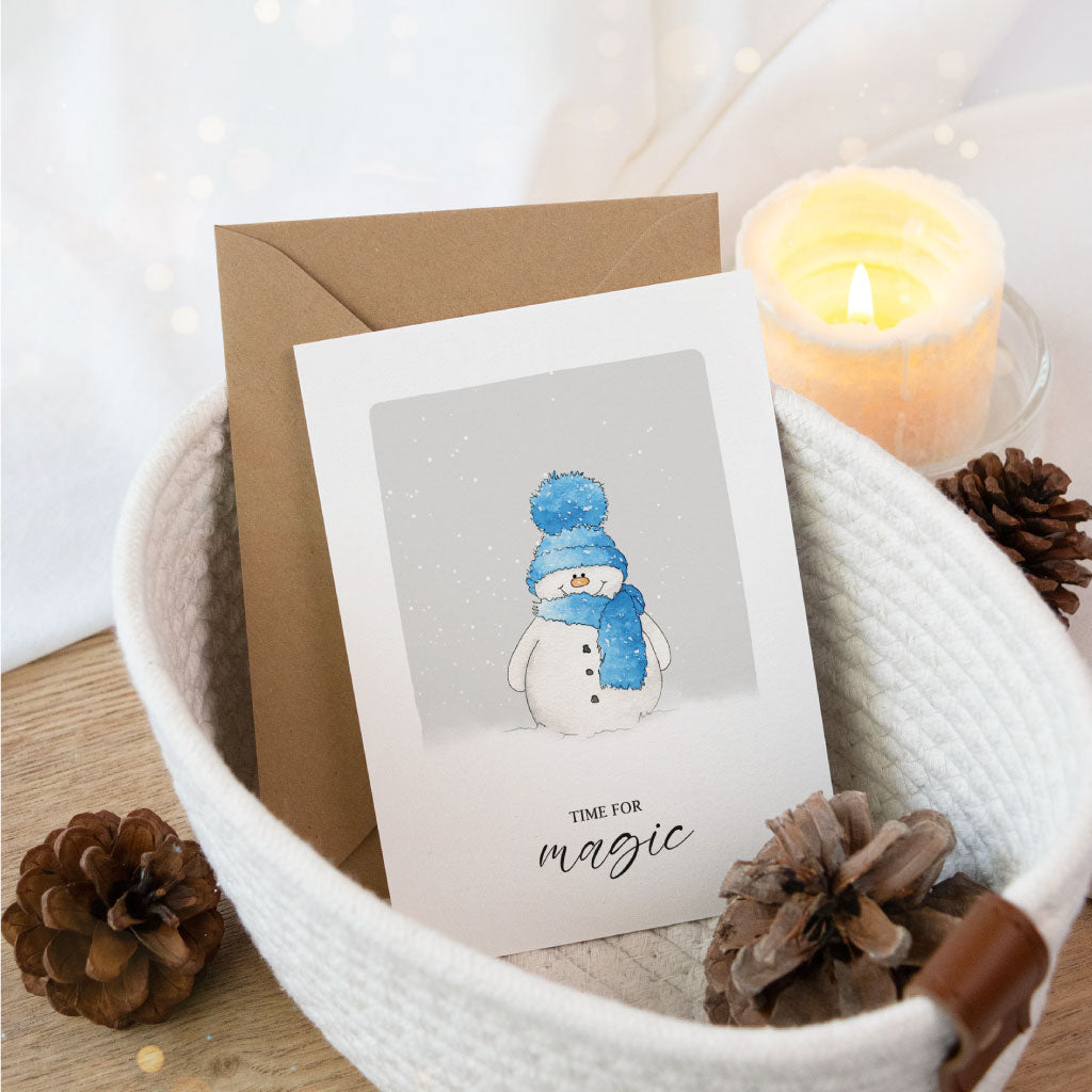 Klappbare Weihnachtskarte mit handgemaltem Schneemann im Aquarellstil, winterliches Motiv mit blauem Schal und Mütze, darunter der Schriftzug „Time for magic“, liebevoll arrangiert mit Umschlag und Deko.