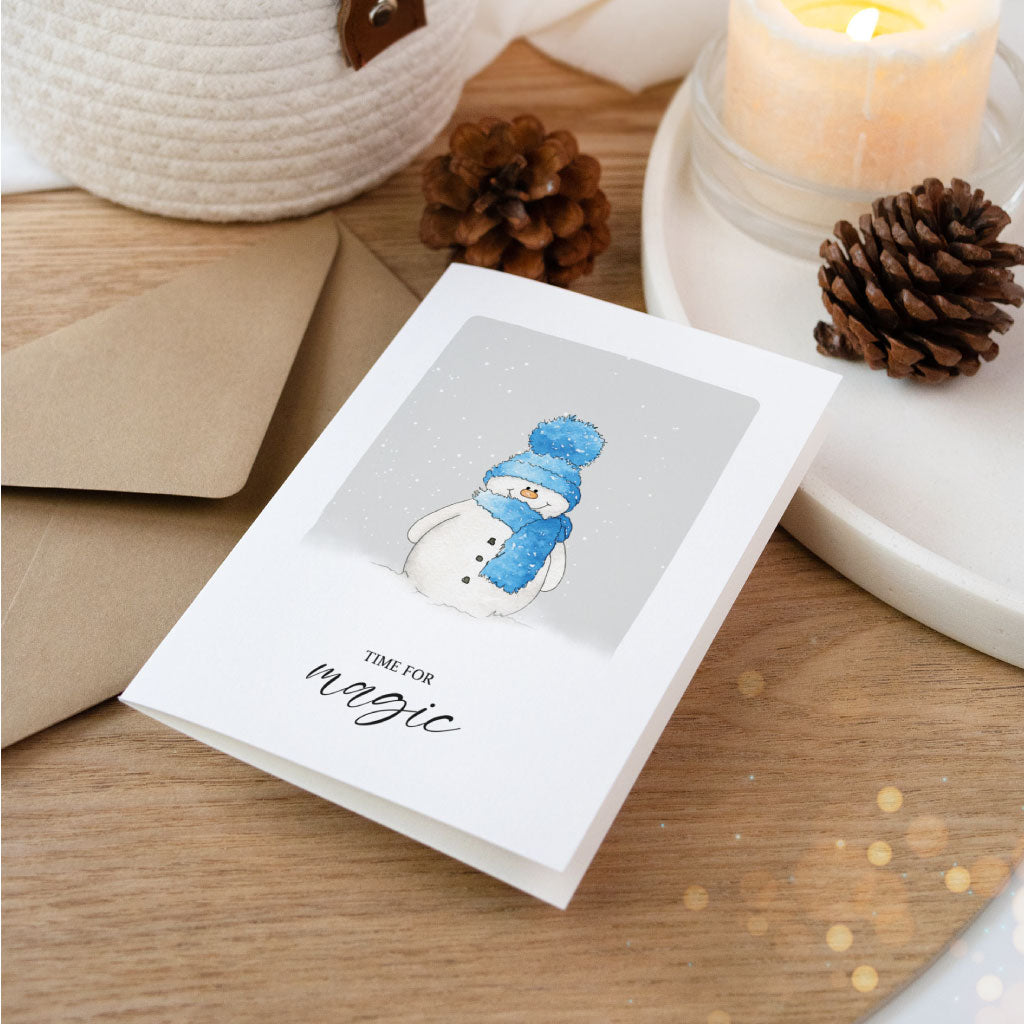 Stimmungsvolle Künstlerkarte mit Schneemann-Aquarell auf Klappkarte, ideal für Weihnachten oder den Winter, daneben ein brauner Umschlag und natürliche Deko.