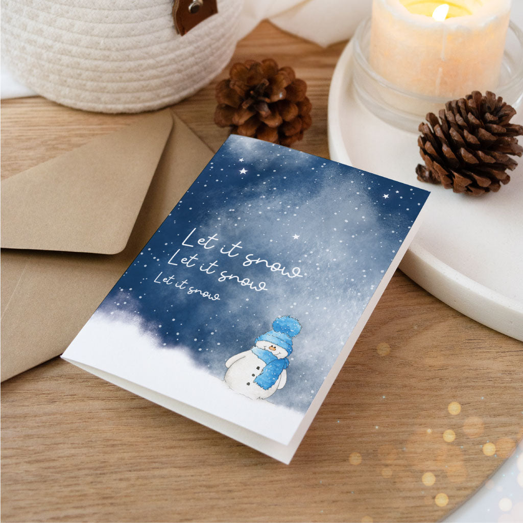 Stimmungsvolle Weihnachtskarte mit handgemaltem Schneemann im Aquarell-Stil und dem Spruch „Let it snow“, neben Tannenzapfen, Kerze und Umschlag arrangiert – ideal zum Weihnachtsgeschenke verpacken.