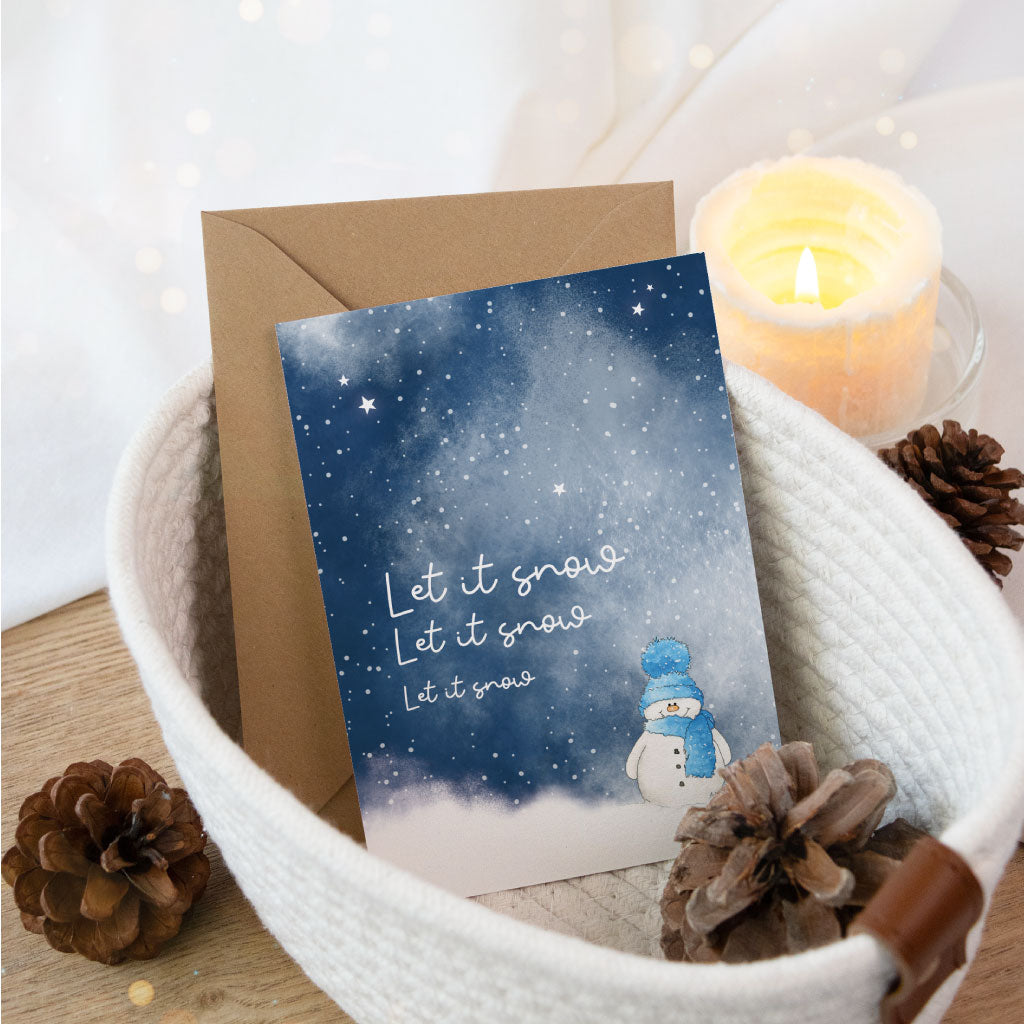 Besondere handgemachte Postkarte mit niedlichem Schneemann-Aquarell und „Let it snow“-Aufschrift, dekorativ in einem Stoffkorb mit Kerze und Tannenzapfen inszeniert.