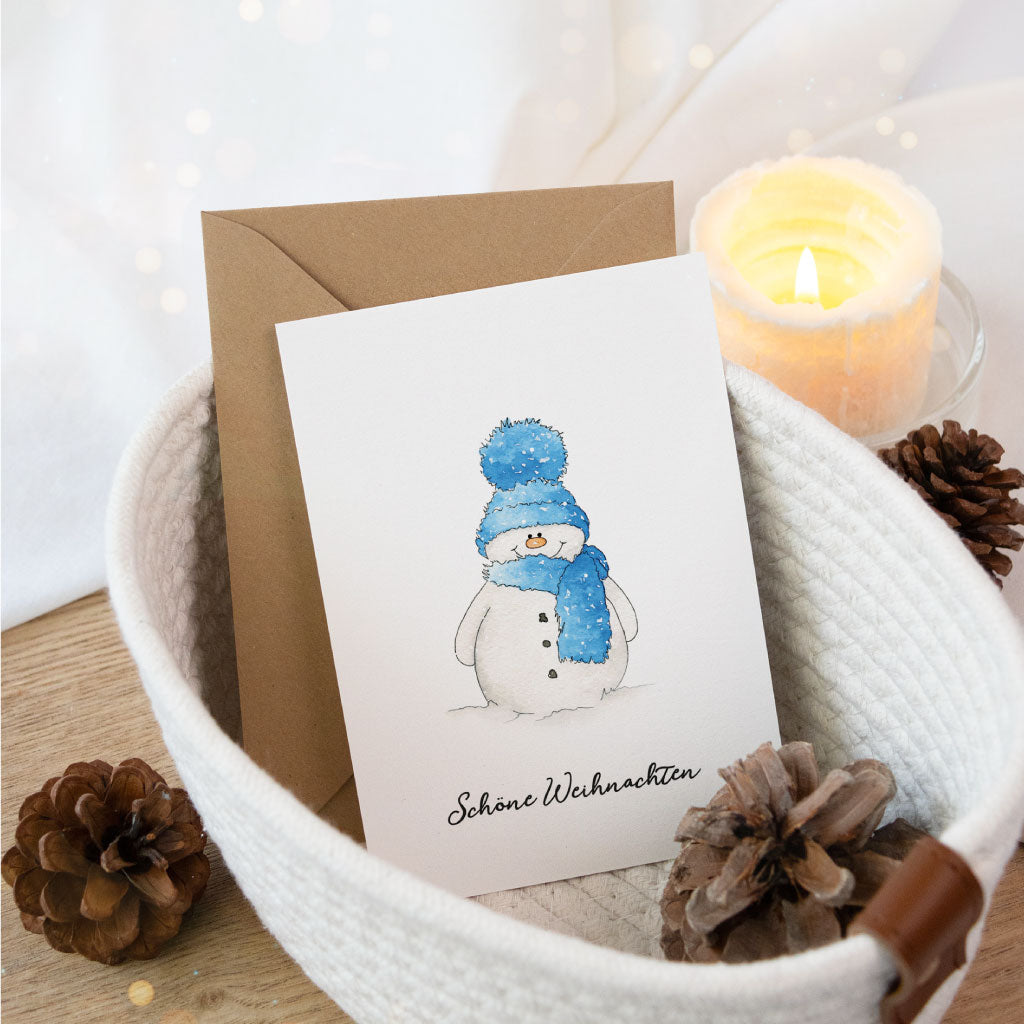 Weihnachtskarte mit handgemaltem Aquarell-Schneemann in blauer Mütze und Schal, festlich dekoriert mit Kerze und Zapfen – perfekte Postkarte zum Weihnachtsgeschenke verpacken.