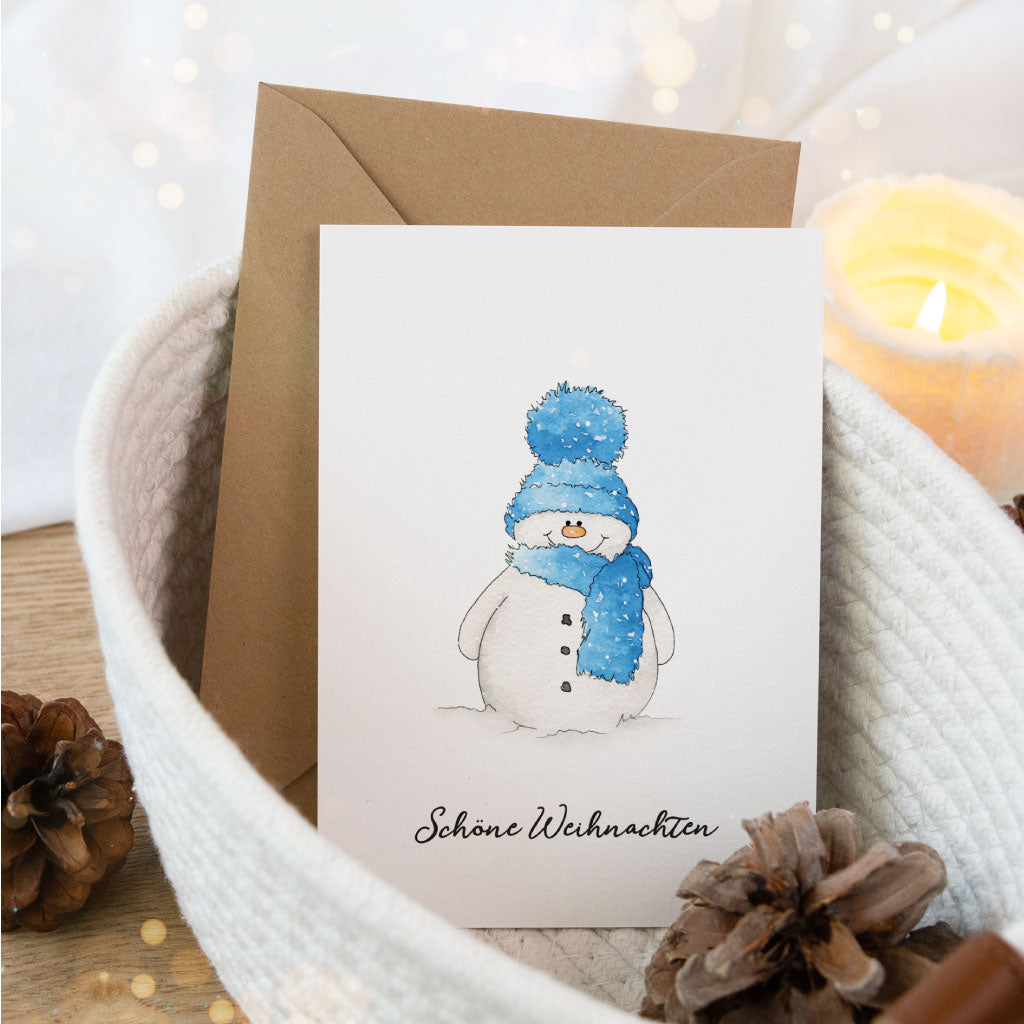 Aquarell-Weihnachtskarte mit Schneemann in blauer Winterkleidung, arrangiert in gemütlicher Weihnachtsdekoration mit Kerze und Tannenzapfen.