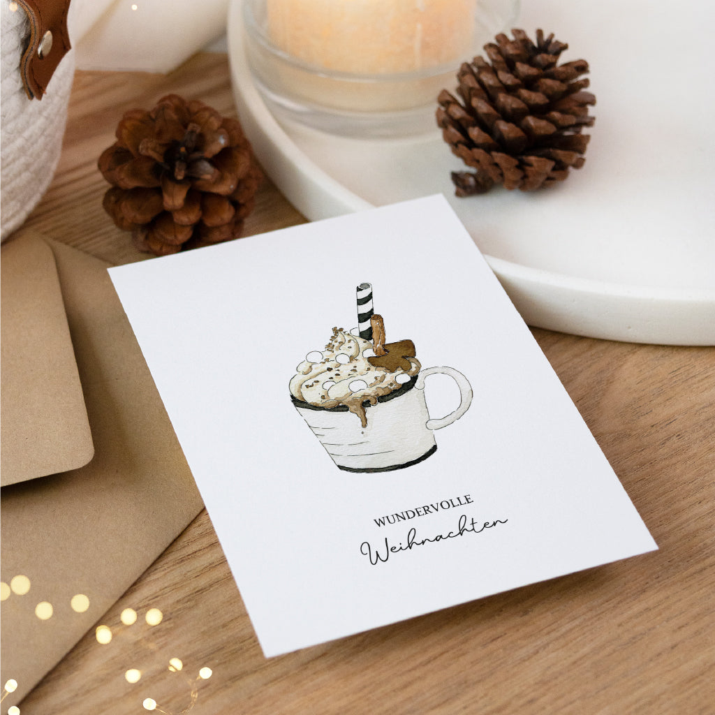 Schräg liegende Weihnachtskarte mit Glühwein Aquarell Motiv neben Umschlag und Tannenzapfen auf rustikalem Holztisch.