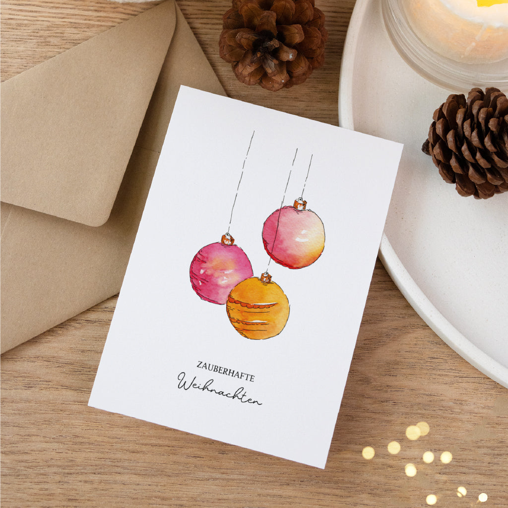Dekorative Postkarte für Weihnachtsgrüße mit rosa und orangen Kugeln als Aquarell Motiv neben Kraftpapier Umschlag.
