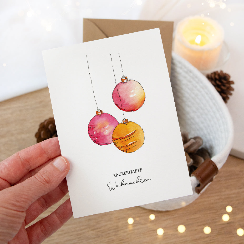 Eine Hand hält die edle Weihnachtskarte mit dem zauberhaften Weihnachtskugeln Aquarell Motiv vor einem weißen Korb.