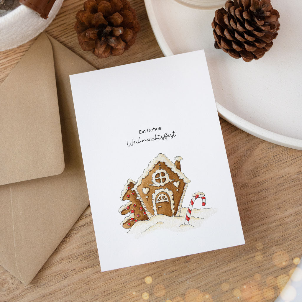 Postkarte mit handgemaltem Aquarell eines Lebkuchenhauses und dem Text „Ein frohes Weihnachtsfest“, festlich arrangiert mit Umschlag und Tannenzapfen.