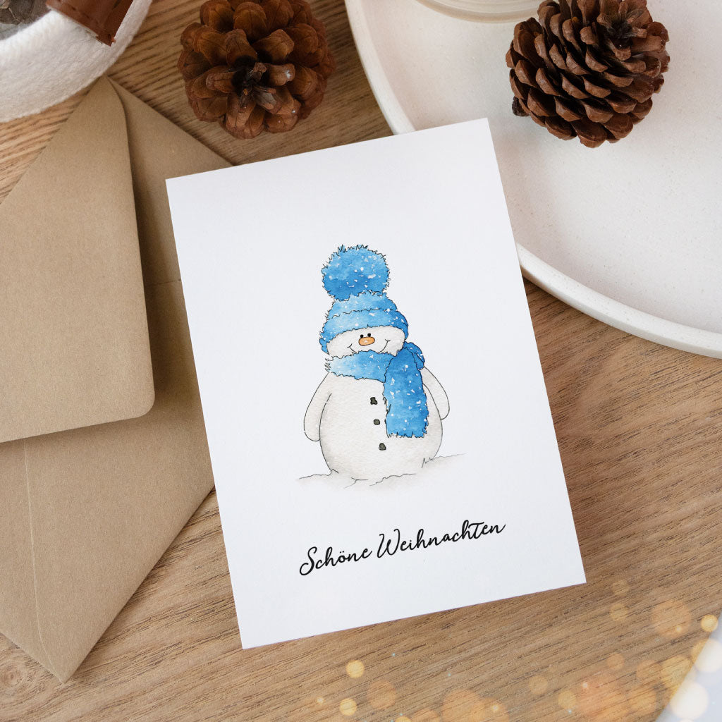 Grußkarte mit Aquarell-Schneemann in Mütze und Schal und dem Text „Schöne Weihnachten“, stimmungsvoll arrangiert mit Kraftpapierumschlag und Deko.